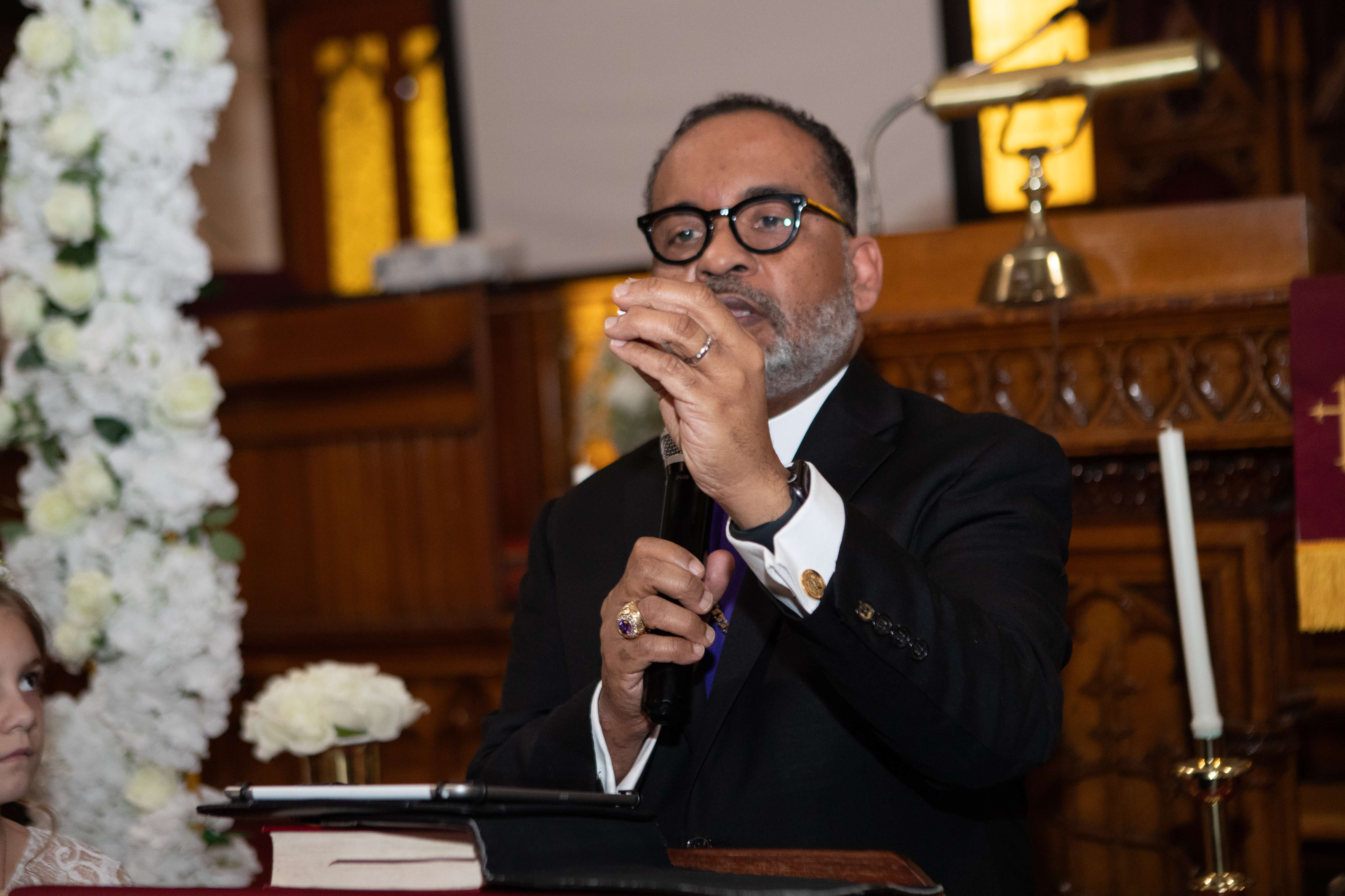 Officiant in a black suit and purple stole speaking into a microphone beside a white floral arch