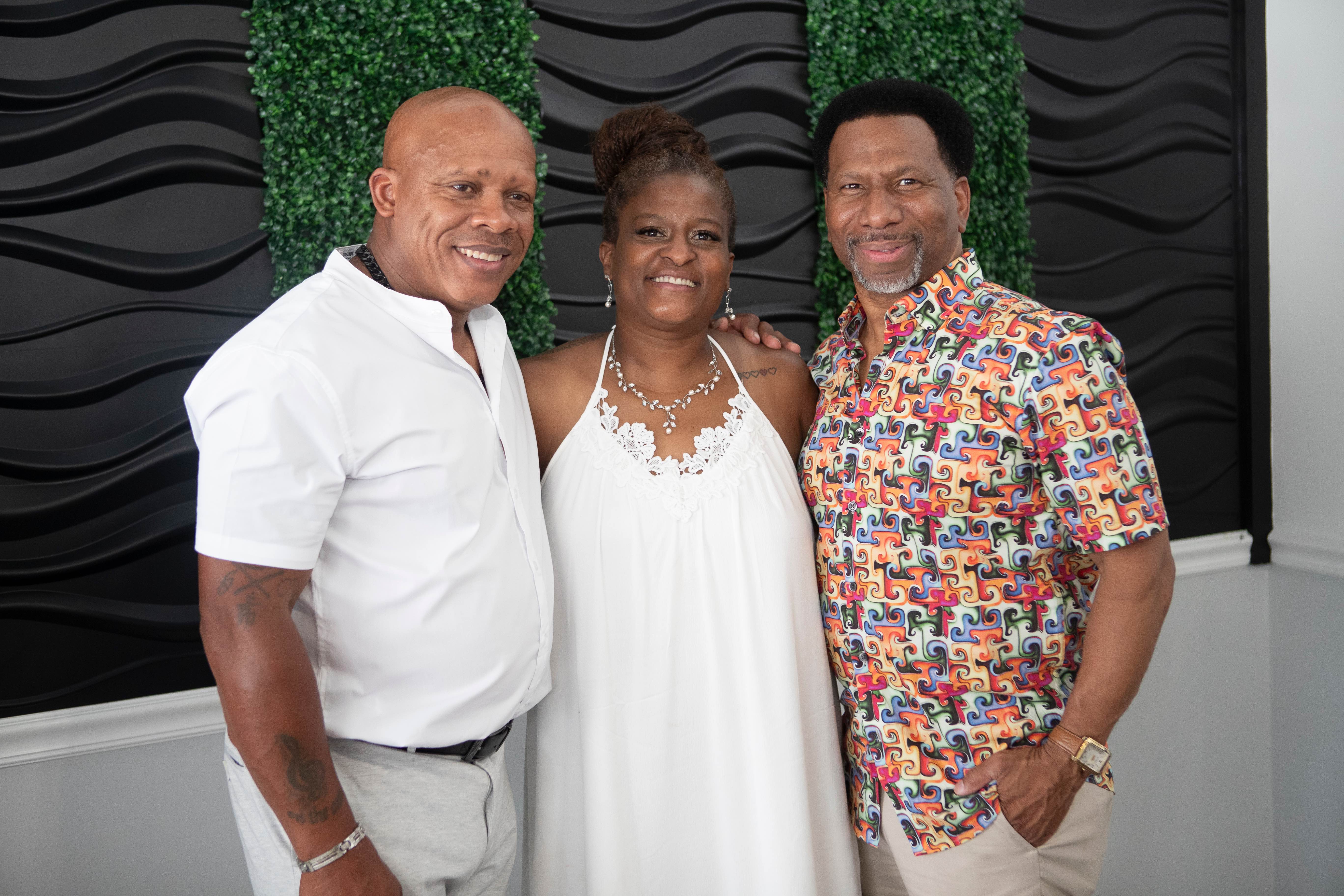 Three guests smiling together in front of a black wave panel and greenery backdrop at a celebration