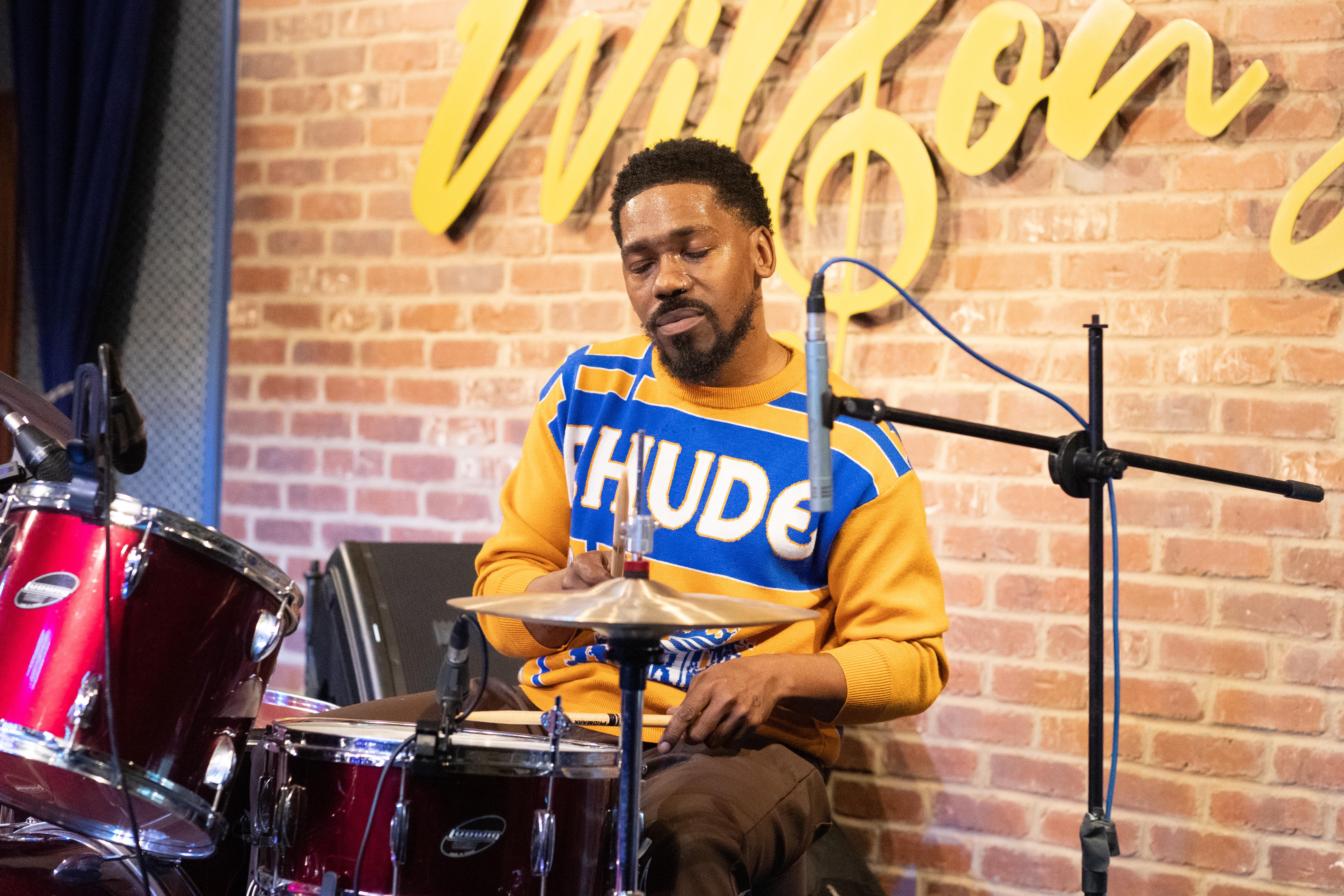 Drummer in a yellow and blue sweater performing on a red kit at a live music venue