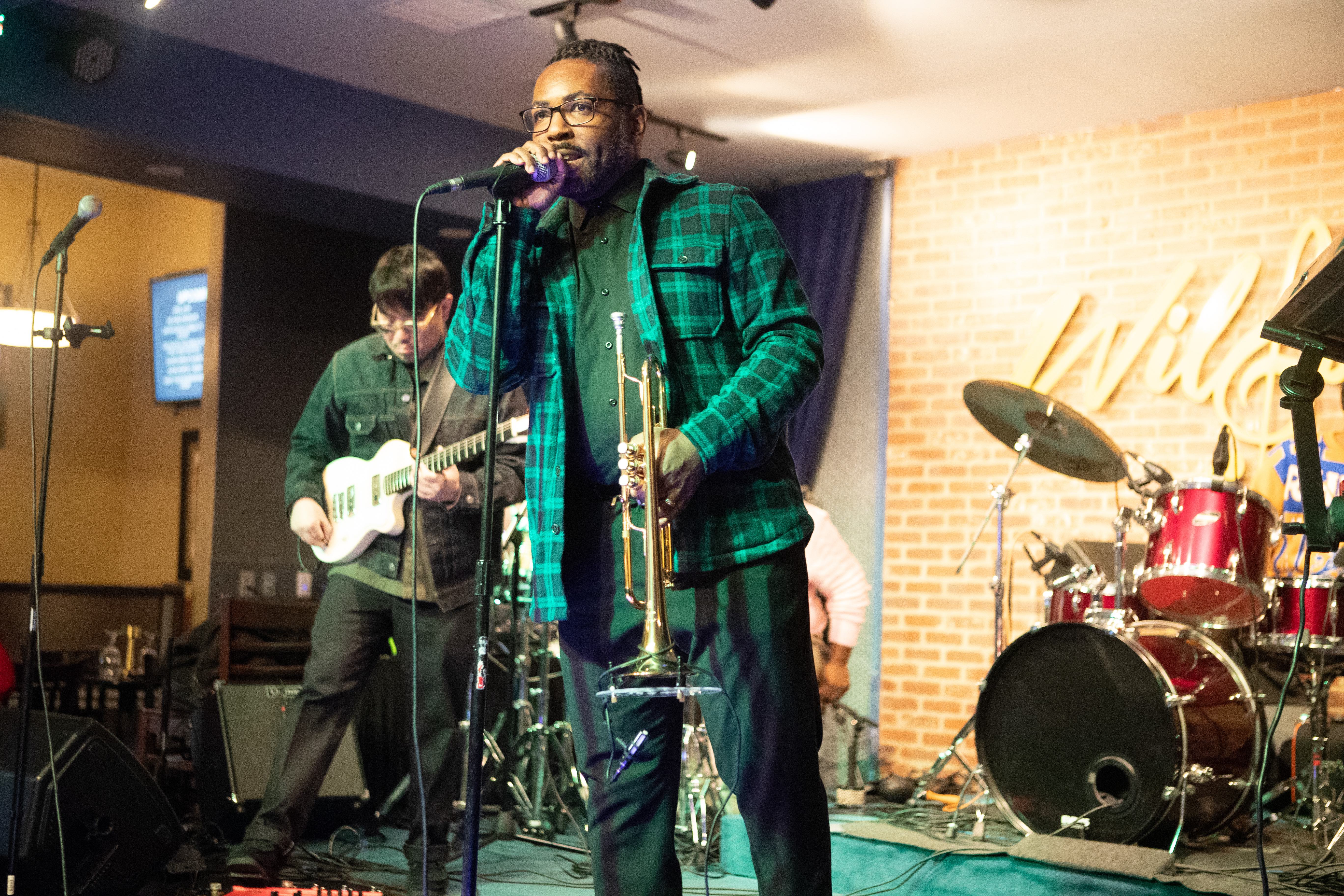 Vocalist in a green plaid jacket singing into a microphone while holding a trumpet, guitarist behind him