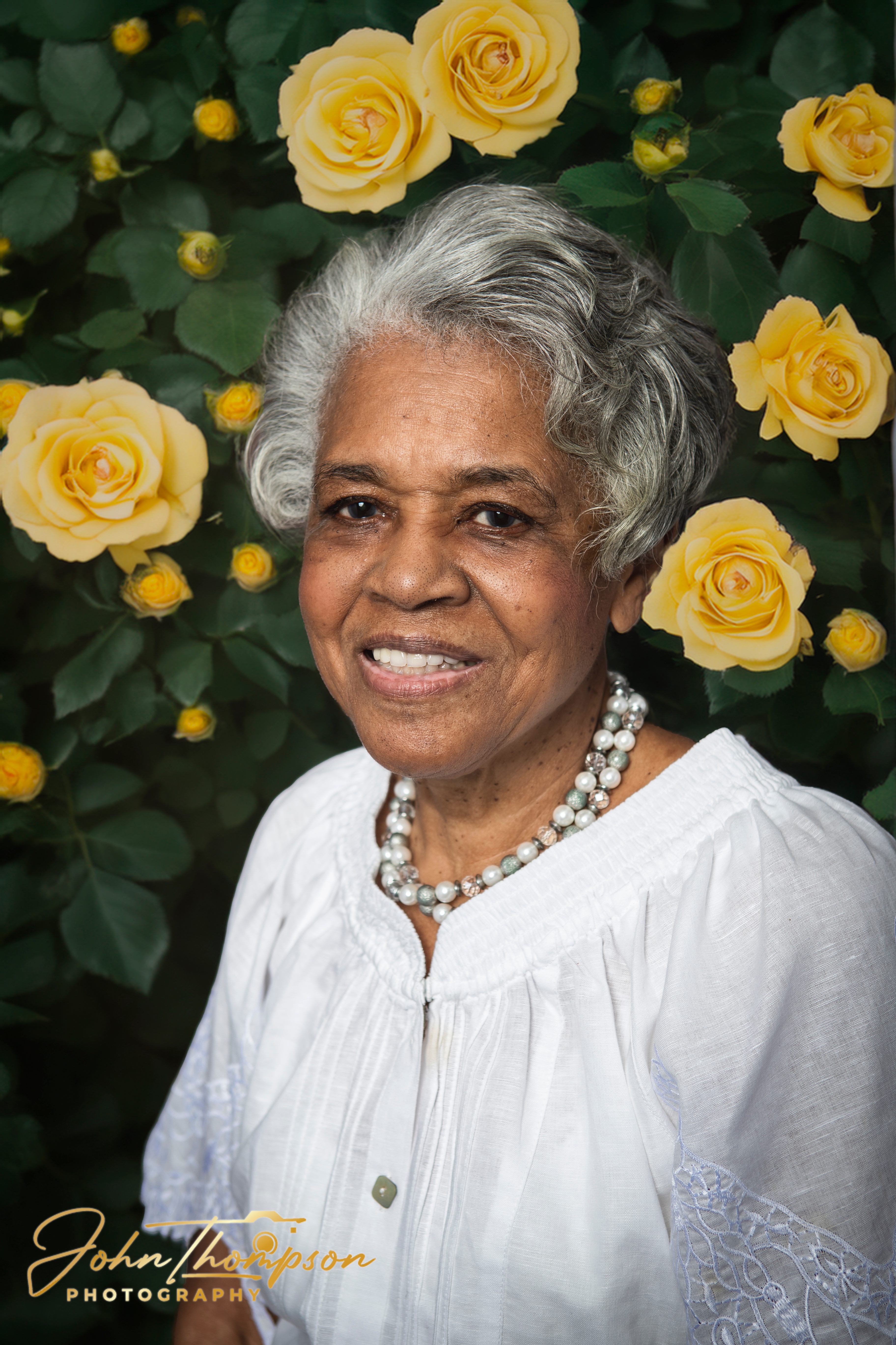 Portrait of a woman with silver hair in a white blouse and pearls standing before a yellow rose bush