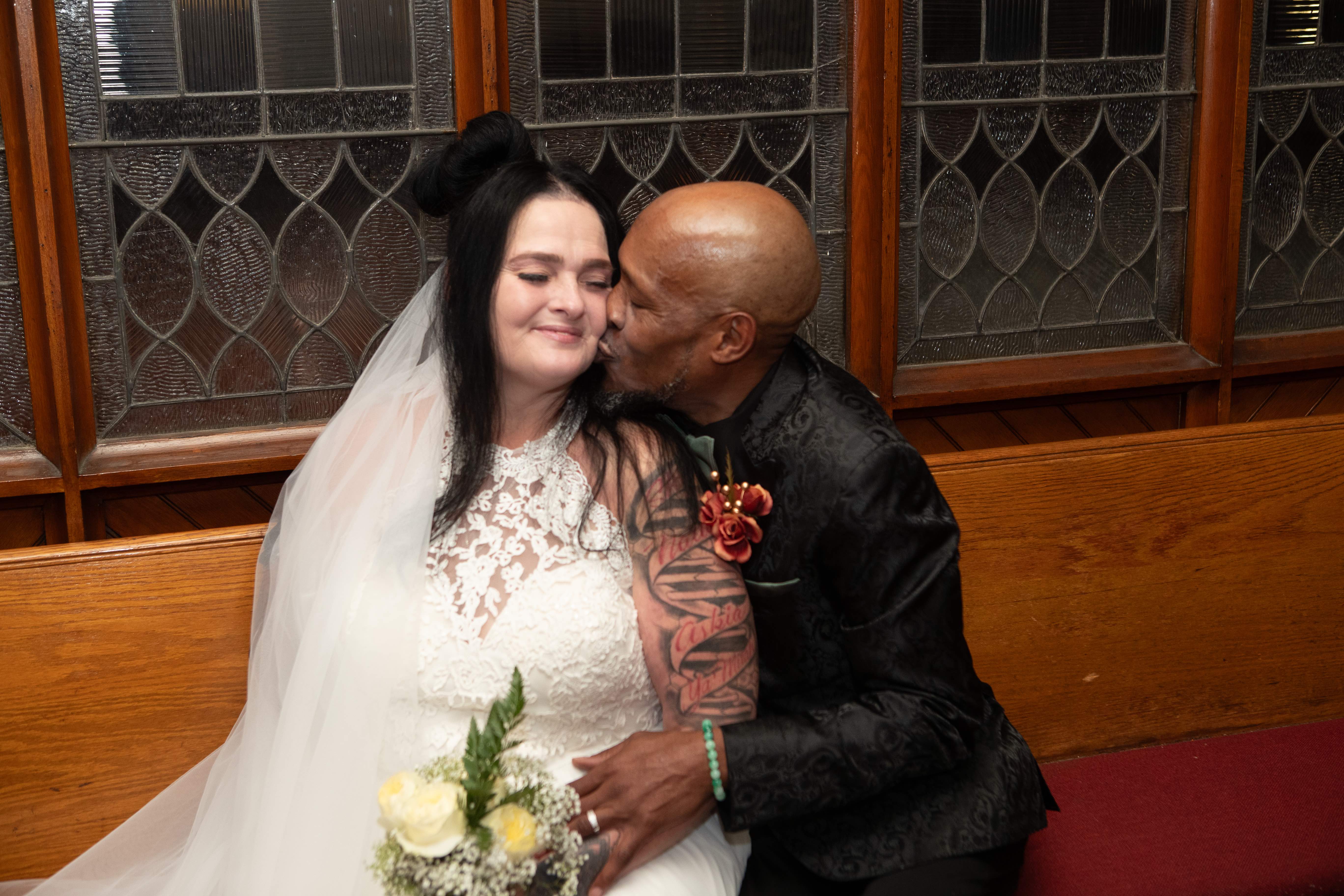Groom kissing his bride on the cheek in a church with stained glass windows behind them