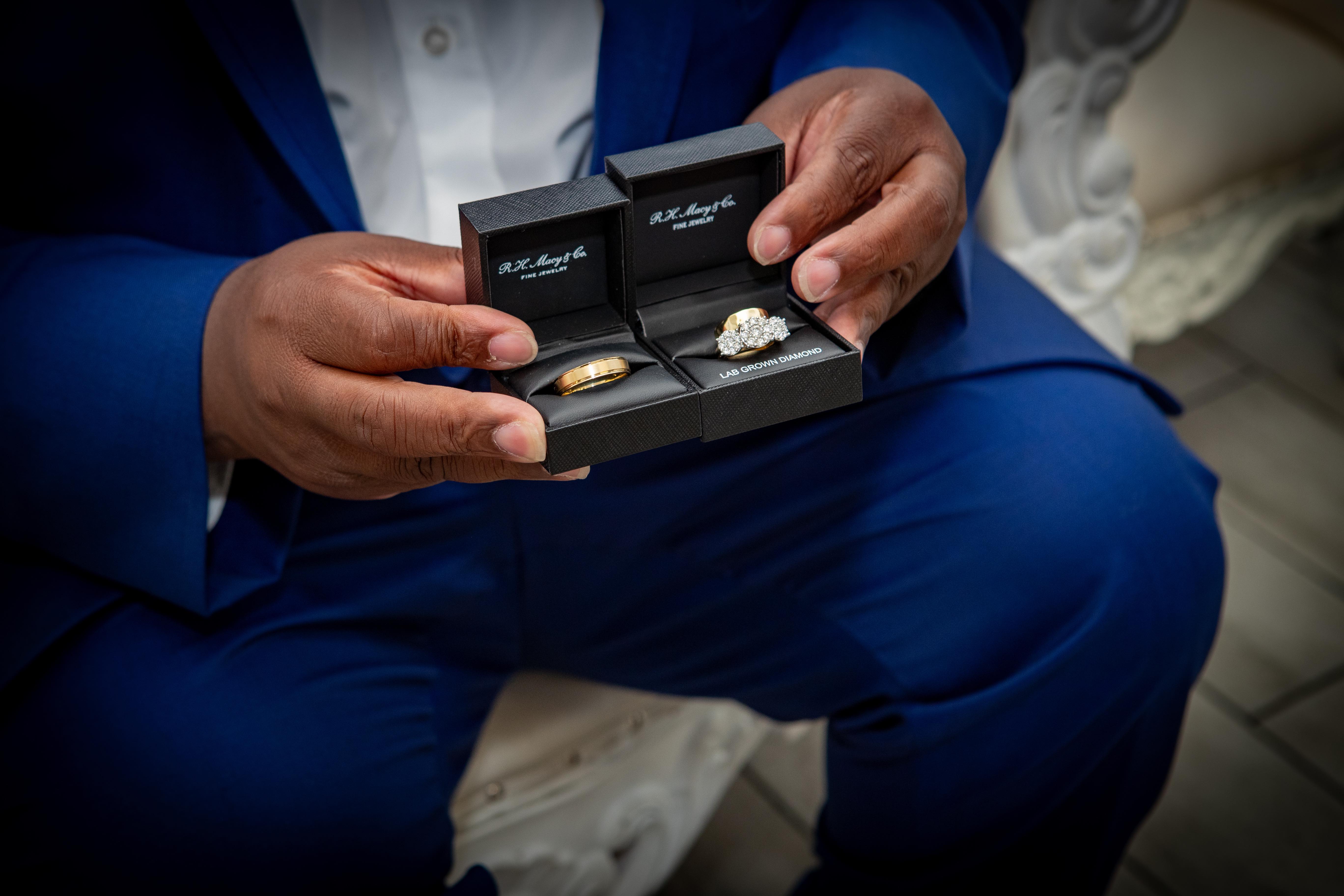 Groom in a royal blue suit holding two open ring boxes with a gold band and a diamond ring